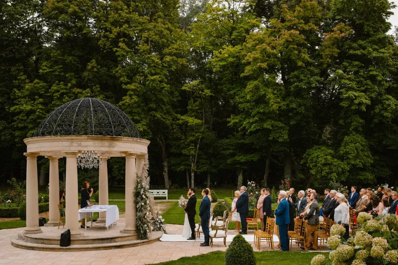Uroczystość zaślubin w altanie, gdzie można wziąć ślub cywilny. Para młoda, goście i urzędniczka w otoczeniu zieleni.