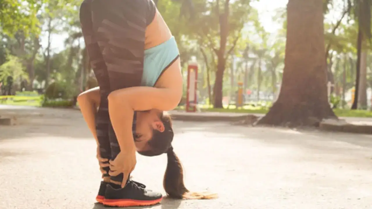 Femme en tenue de sport effectuant un étirement abdos en se penchant en avant, mains agrippant ses chevilles, dans un parc ensoleillé.