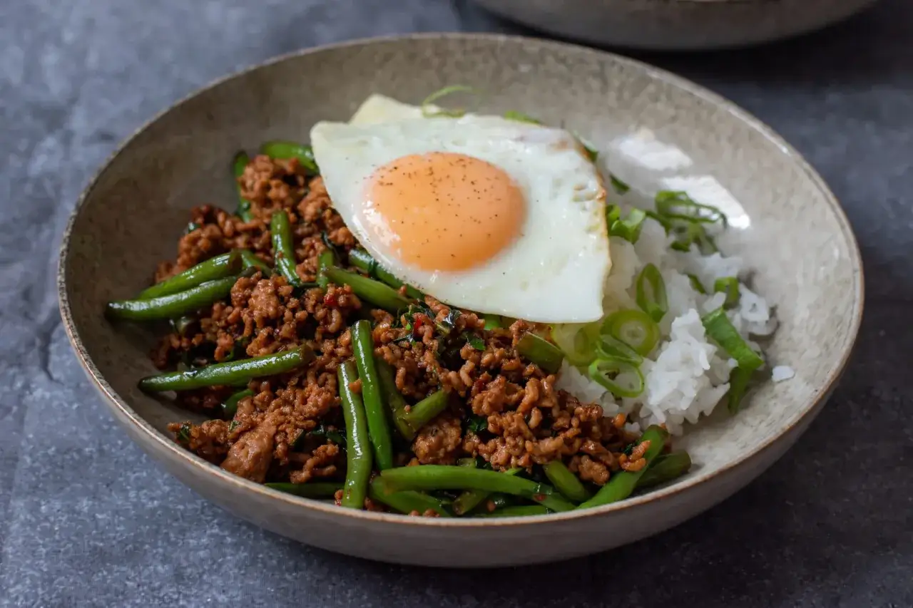 En skål med ris, strimlat kött, gröna bönor och ett stekt ägg. En smakrik pad kra pao som ser utsökt ut.