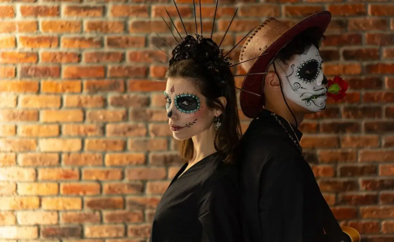 Para stojąca plecami do siebie, ubrana w kostiumy inspirowane Día de Muertos. Kobieta ma makijaż czaszki i ozdobę głowy, mężczyzna kapelusz i róż w ustach.
