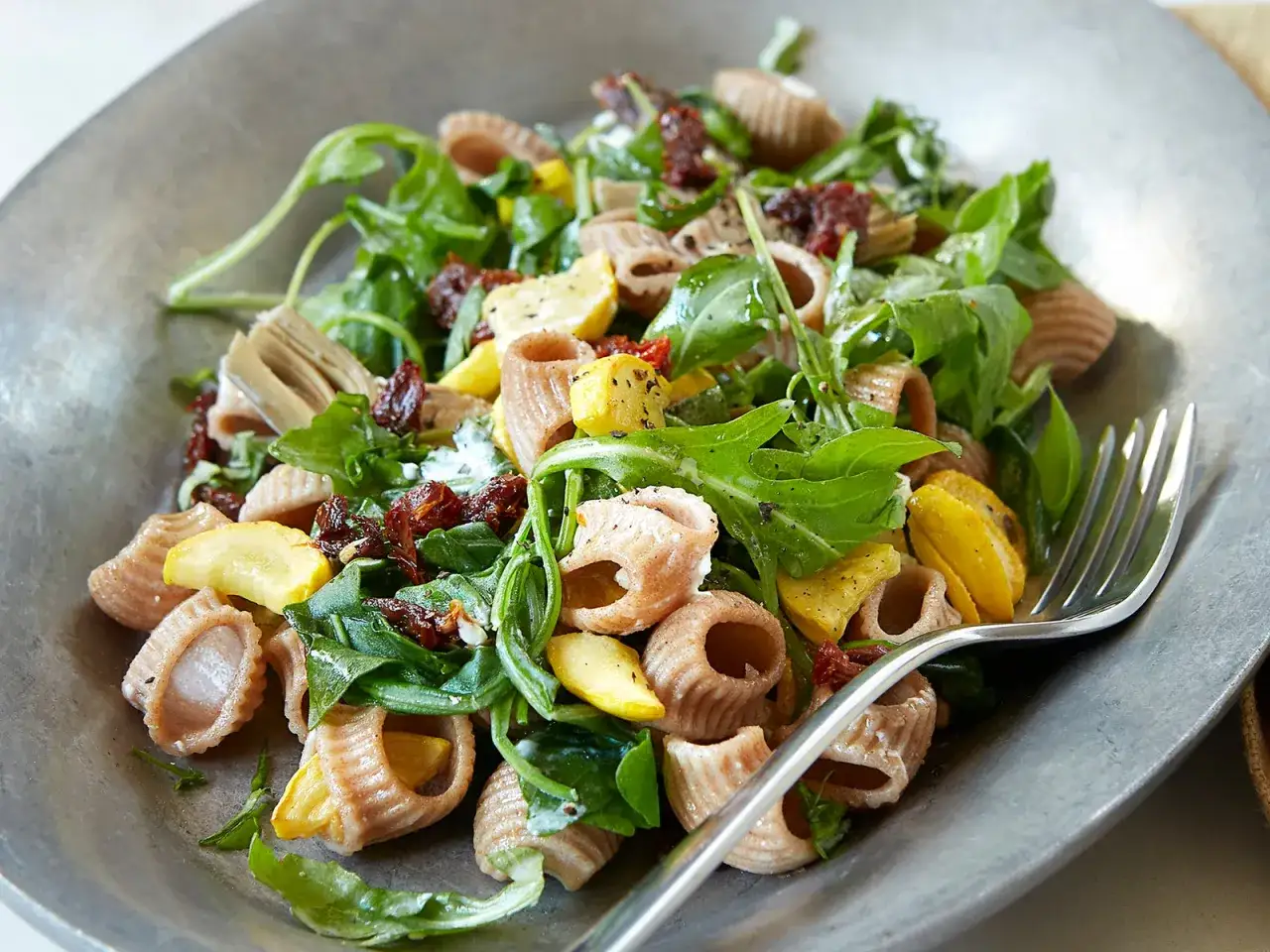 Insalata di pasta integrale con rucola, zucchine, pomodori secchi e carciofi. Un piatto gustoso e leggero, ideale per chi segue un deficit calorico.