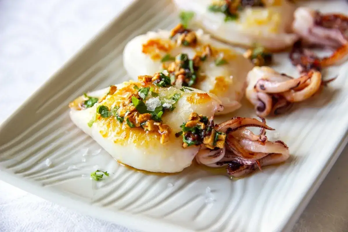 Chipirones a la plancha, tiernos y dorados, con un toque de ajo y perejil picado, servidos en una elegante bandeja blanca.