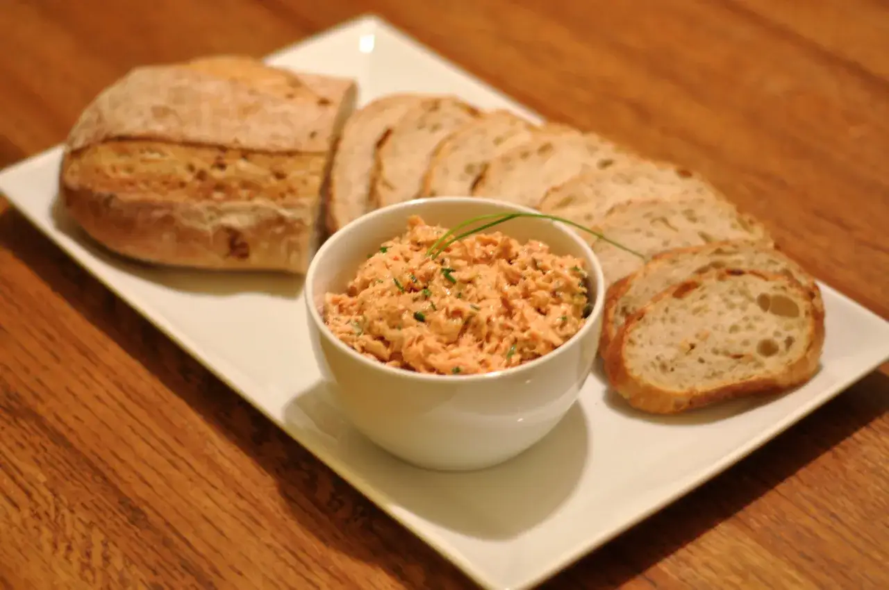 Un bol de rillettes de saumon, garni de ciboulette, servi avec du pain artisanal tranché sur une assiette blanche.