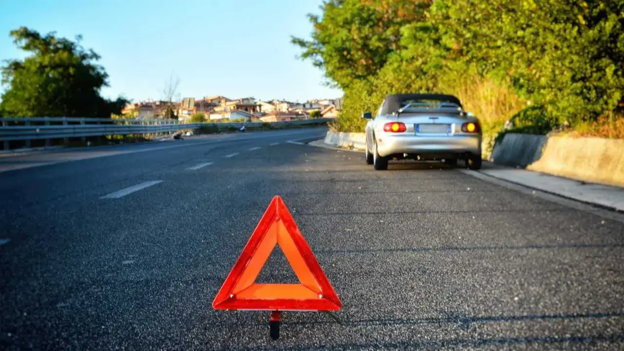 Triangolo rosso di emergenza sulla strada, con un'auto sportiva argentata ferma in lontananza. Le 4 frecce lampeggiano, segnalando un problema.