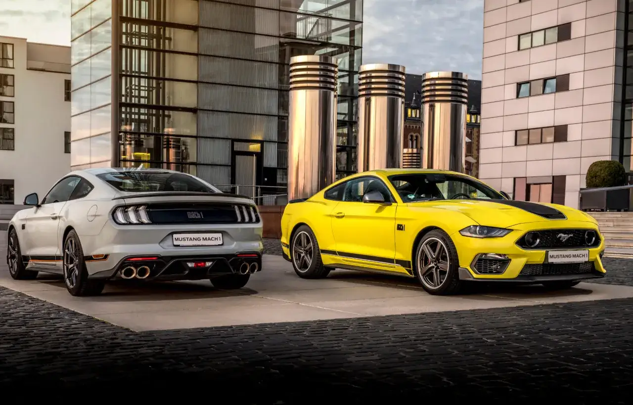Dwa Fordy Mustang Mach 1, jeden żółty, drugi biały, stoją przed nowoczesnym budynkiem.