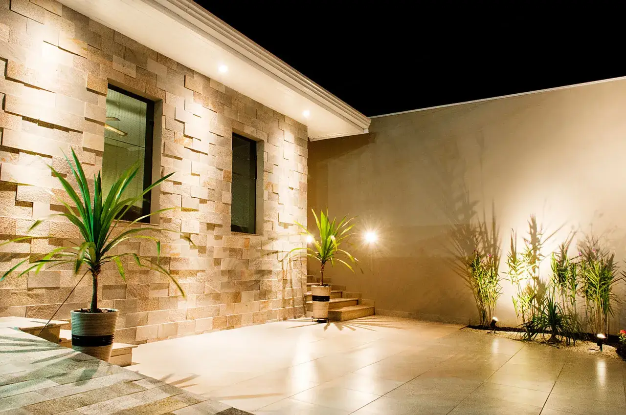 Un cortile illuminato di notte con facciate in pietra e mattoni, piante in vaso e un'area pavimentata.