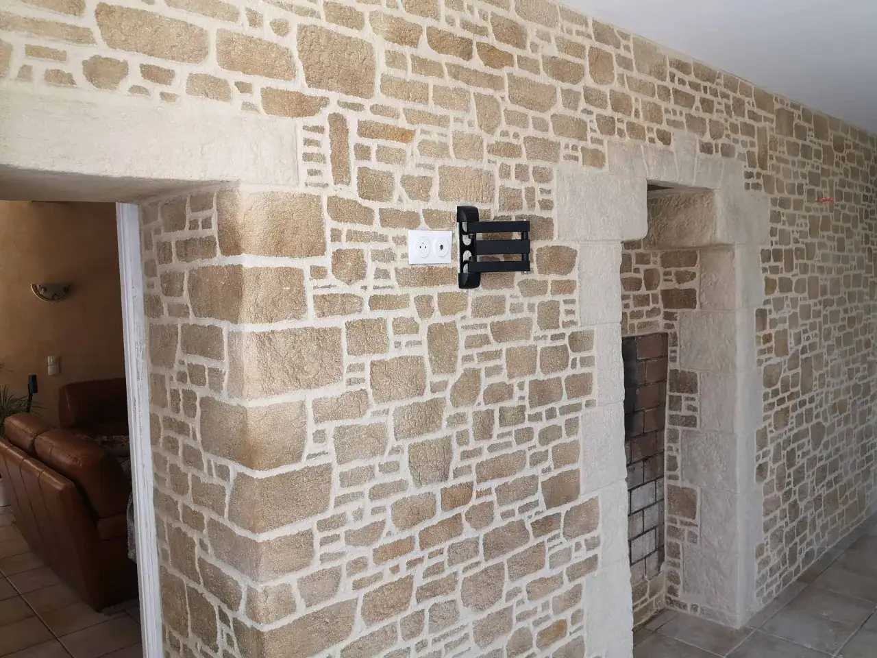 Un mur en **enduit pierre apparente** imitant une ma&ccedil;onnerie ancienne, avec des pierres de tailles irr&eacute;guli&egrave;res et des joints blancs. Un support TV est fix&eacute; au mur.