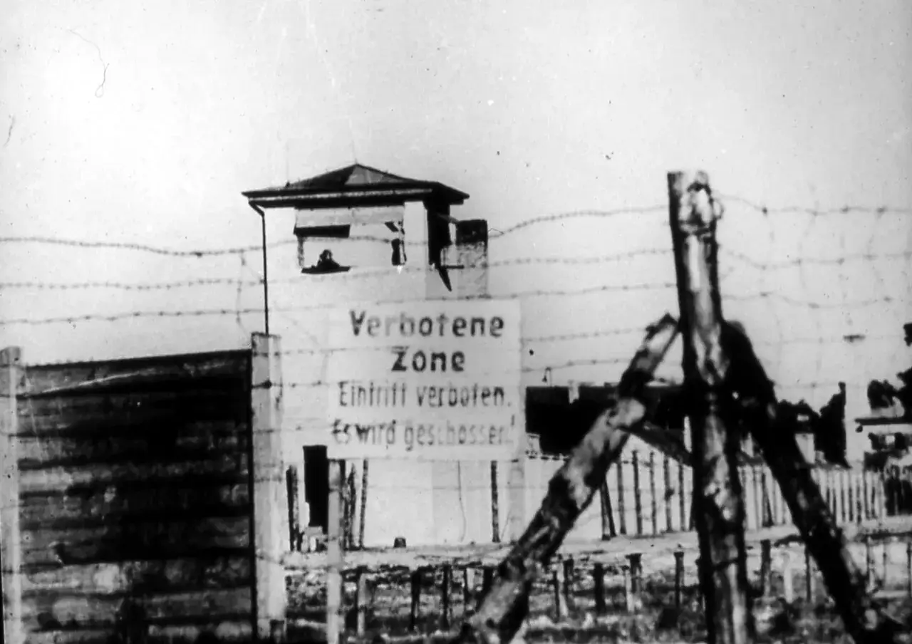Wachturm und Stacheldraht vor einem Tor mit Schild: