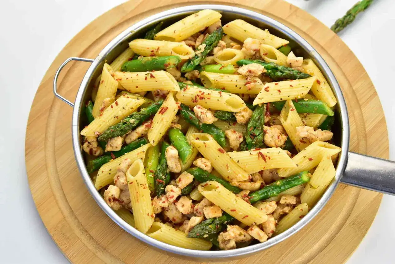 Penne z kurczakiem i zielonymi szparagami, posypane płatkami chili, w metalowej patelni na drewnianej desce.