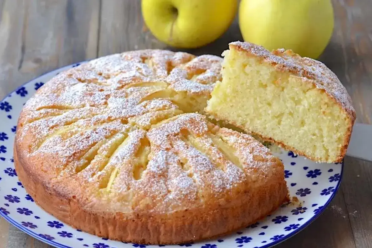 Torta allo yogurt soffice e profumata, decorata con mele e zucchero a velo. Una fetta è già pronta per essere gustata.