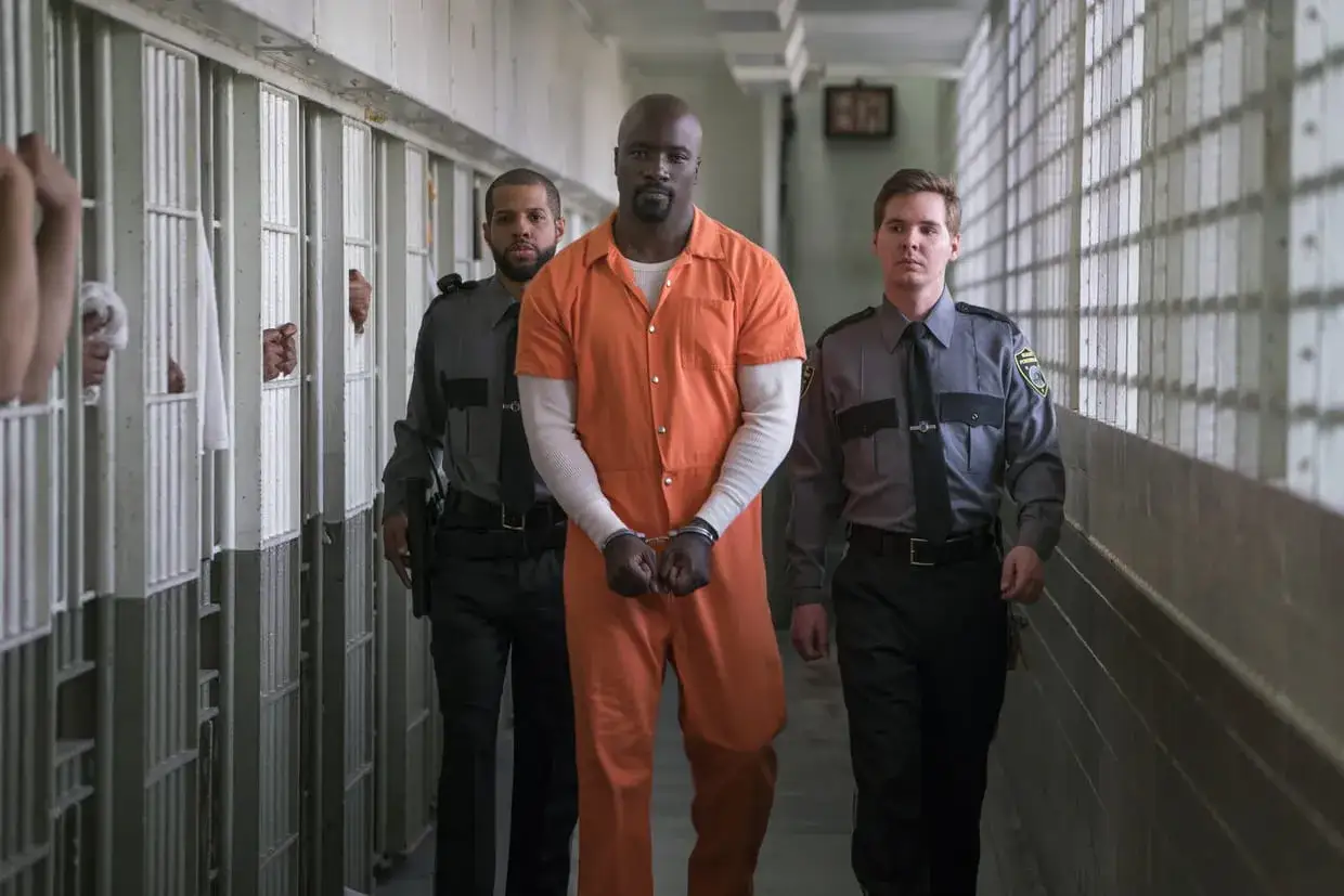 Mike Colter en tenue orange, menotté, escorté par deux gardiens dans un couloir de prison. Scène de films et séries tv avec Mike Colter.