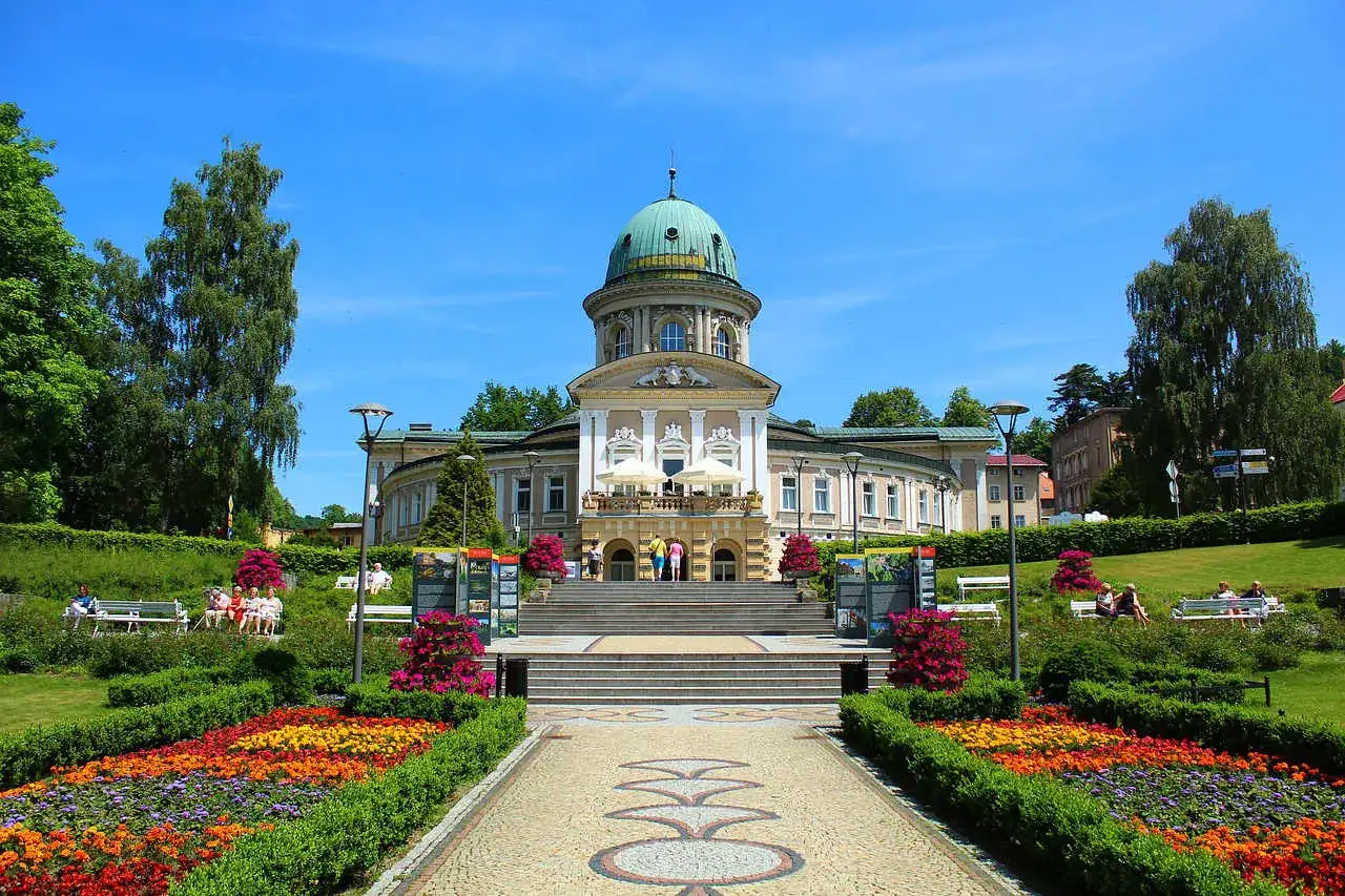 Piękny **ładek zdroj** z kopułą, otoczony kwitnącymi rabatami i zielenią.