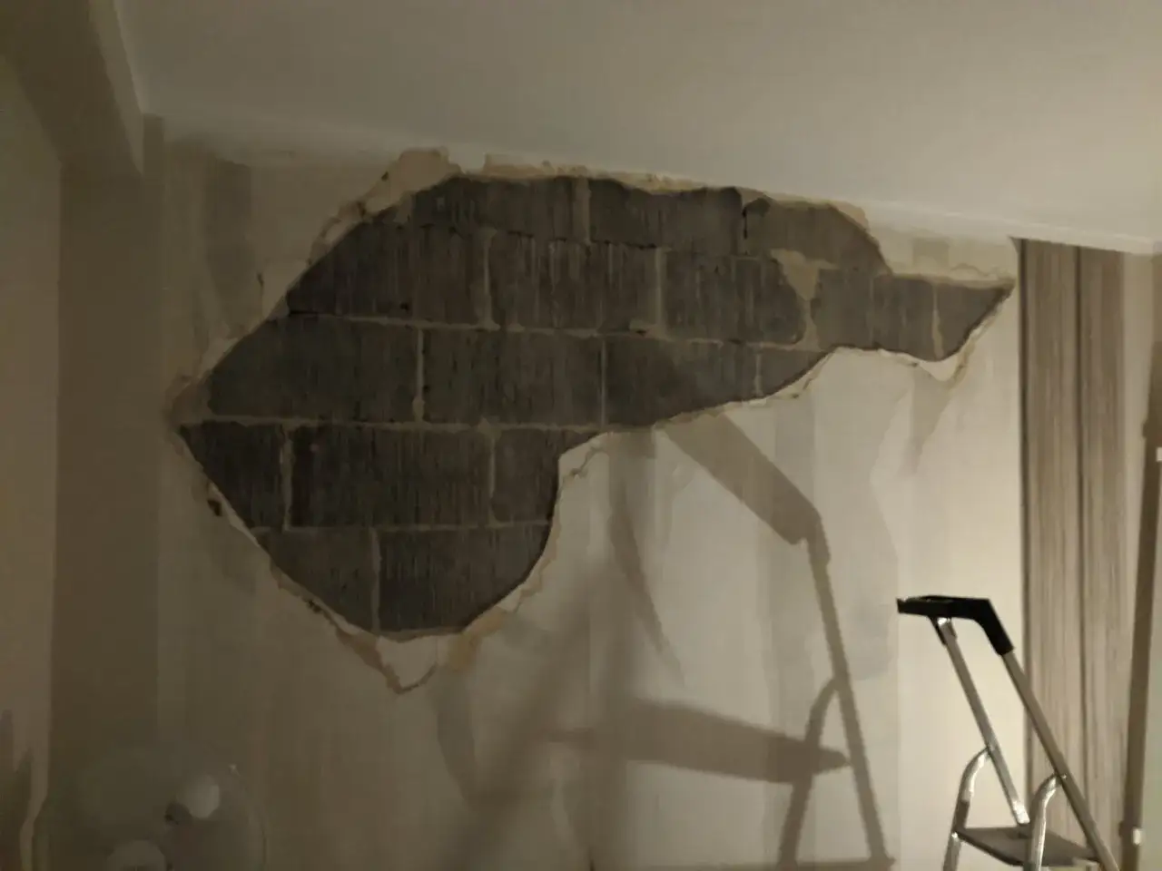 En stor del av tapeten har lossnat och blottar en tegelv&auml;gg. En stege st&aring;r redo f&ouml;r att laga puts inomhus.