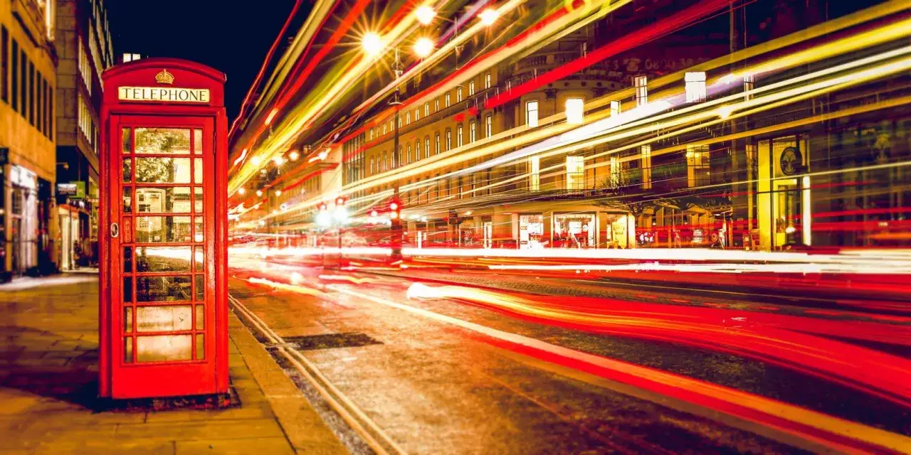 Cabina telefonica rossa iconica, con scie luminose di traffico notturno che sembrano disegnare con la luce la strada e gli edifici circostanti.