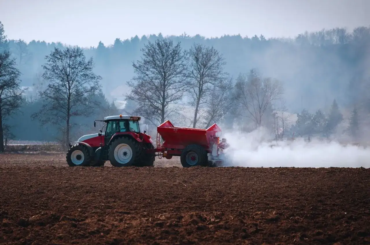 Czerwony traktor z przyczepą rozsypuje nawóz na polu. Wapnowanie gleby jest kluczowe dla wielu warzyw, które lubią wapnowanie, zapewniając im lepszy wzrost.