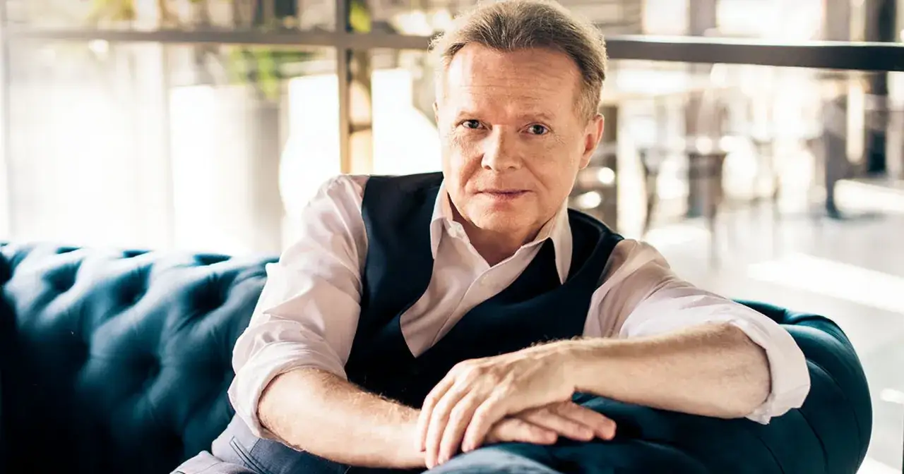 Michał Bajor, w eleganckim stroju, siedzi na granatowej kanapie. Choć na zdjęciu nie ma jego żony, jego spojrzenie sugeruje spokój i pewność siebie.