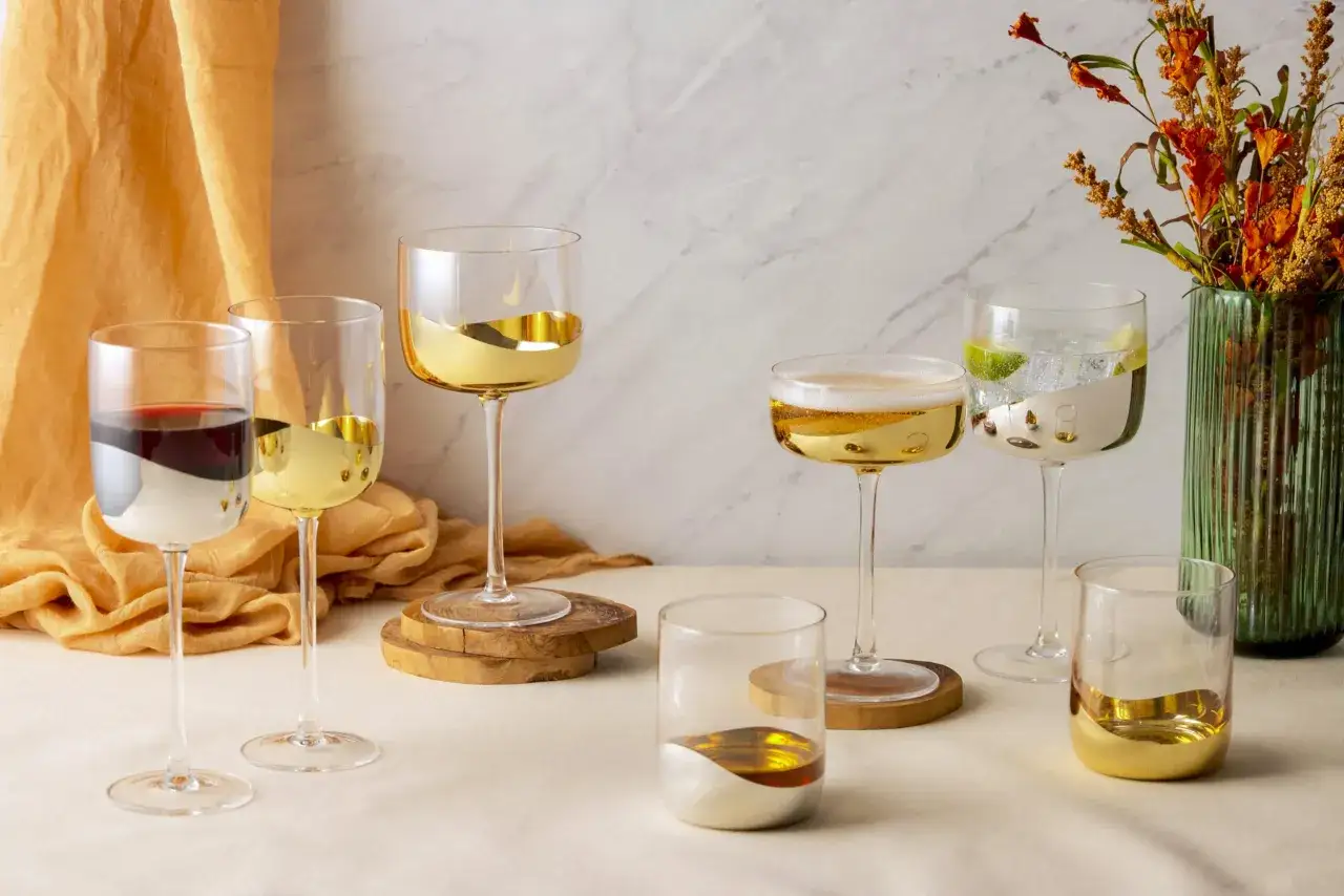 Variedad de tipos de copas de champagne y vasos con detalles dorados y plateados, junto a un jarr&oacute;n con flores secas.
