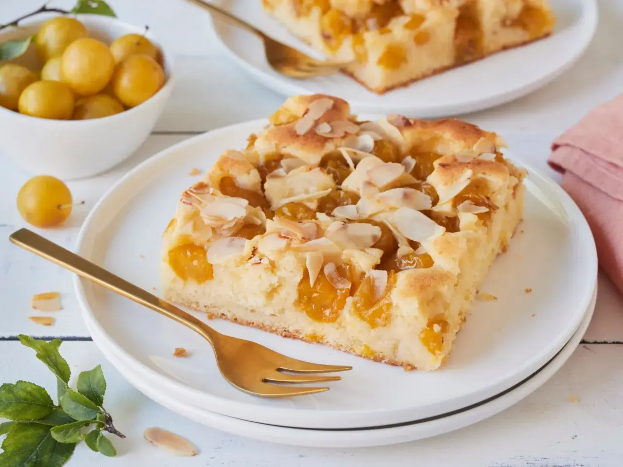 Ein saftiges St&uuml;ck Mirabellenkuchen mit Mandelsplittern auf einem Teller, daneben eine Schale voller frischer Mirabellen.