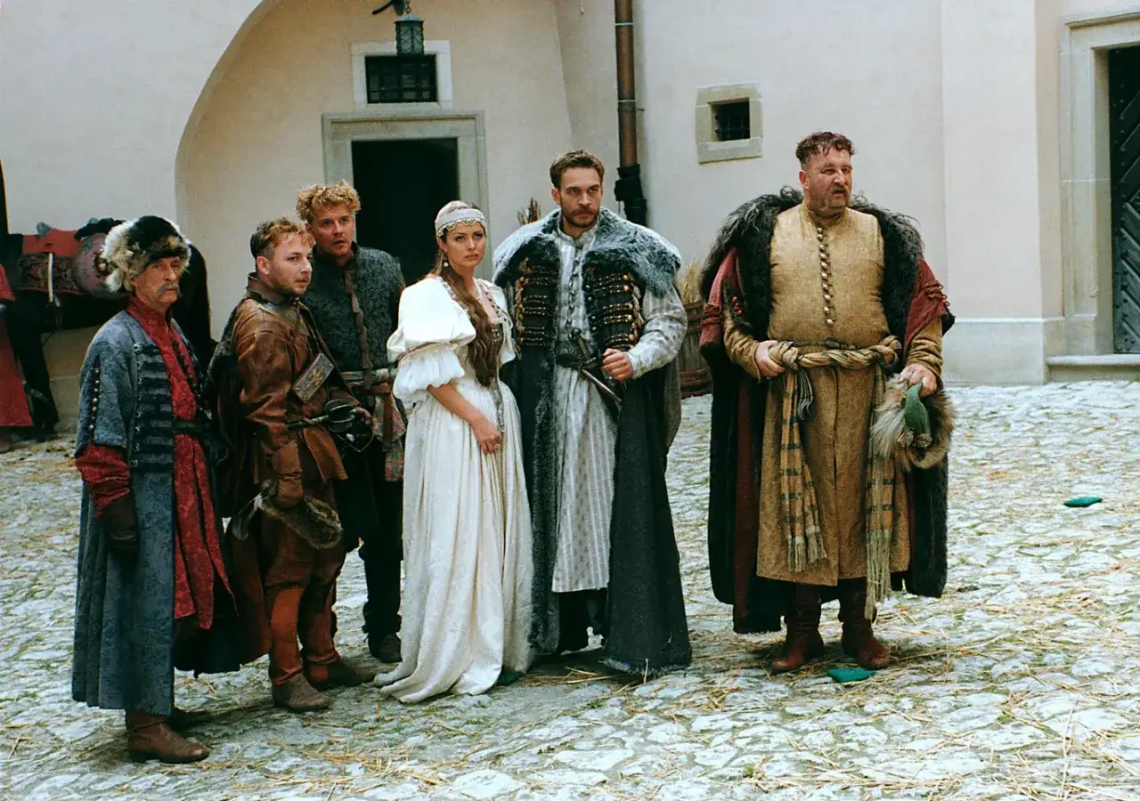 Ogniem i mieczem obsada w historycznych strojach. Grupa postaci, w tym mężczyźni w bogatych szatach i kobieta w białej sukni, stoi na kamiennym dziedzińcu.