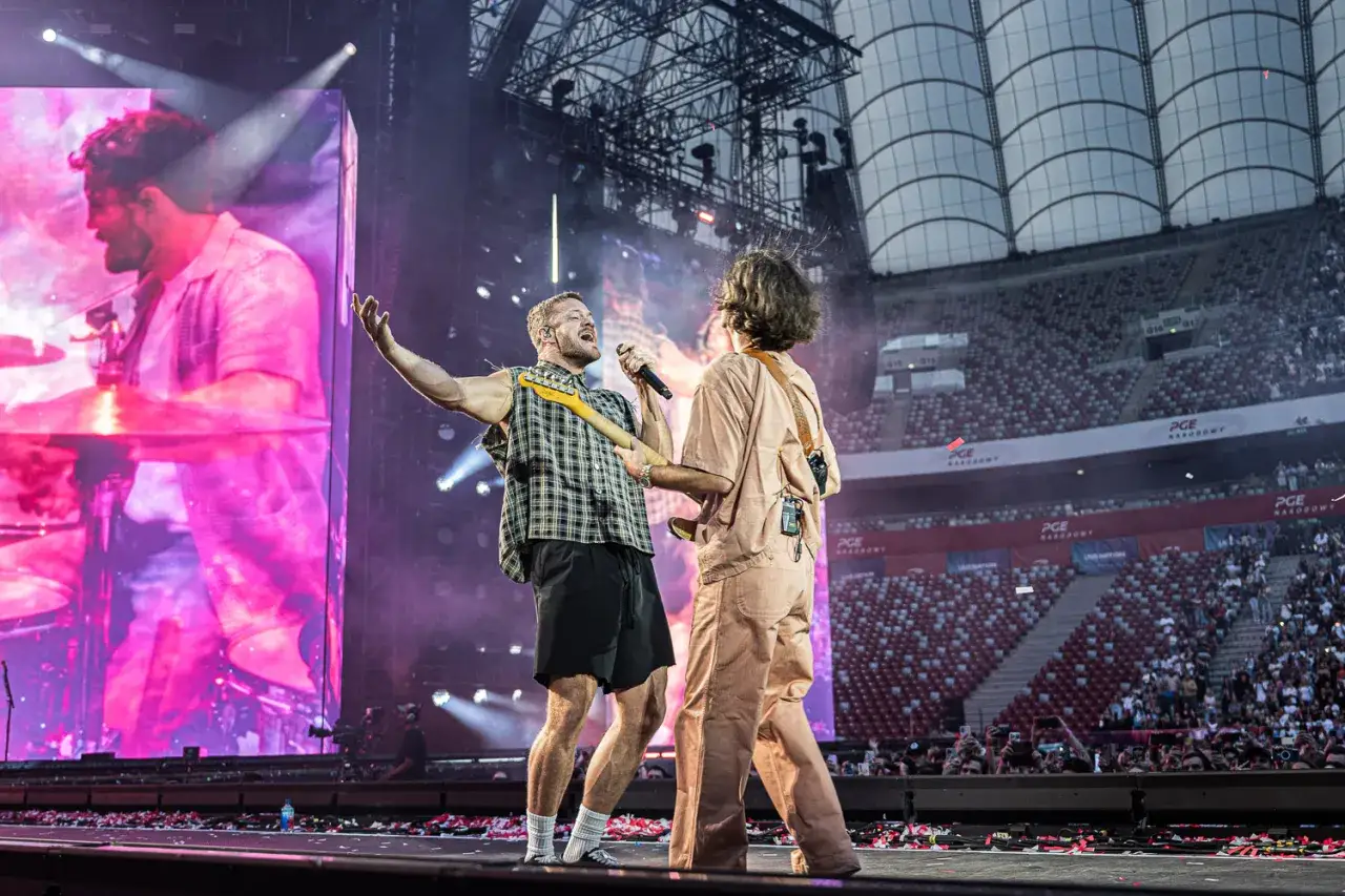 Dan Reynolds z Imagine Dragons śpiewa na koncercie, a obok niego stoi gitarzysta. Tłum na stadionie PGE Narodowy szaleje.