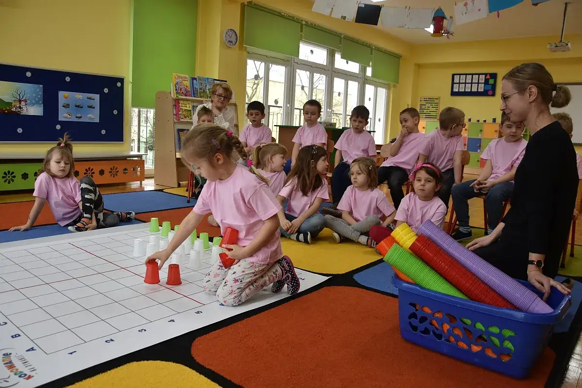 Dzieci w różowych koszulkach bawią się kubeczkami na dywanie. Czy można przenieść dziecko do innej grupy bez zgody rodziców? Nauczycielka obserwuje.