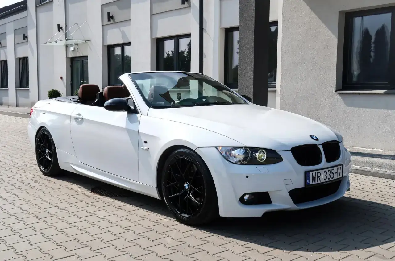 Białe bmw e93 cabrio z brązowym wnętrzem i czarnymi felgami stoi na kostce brukowej przed nowoczesnym budynkiem.