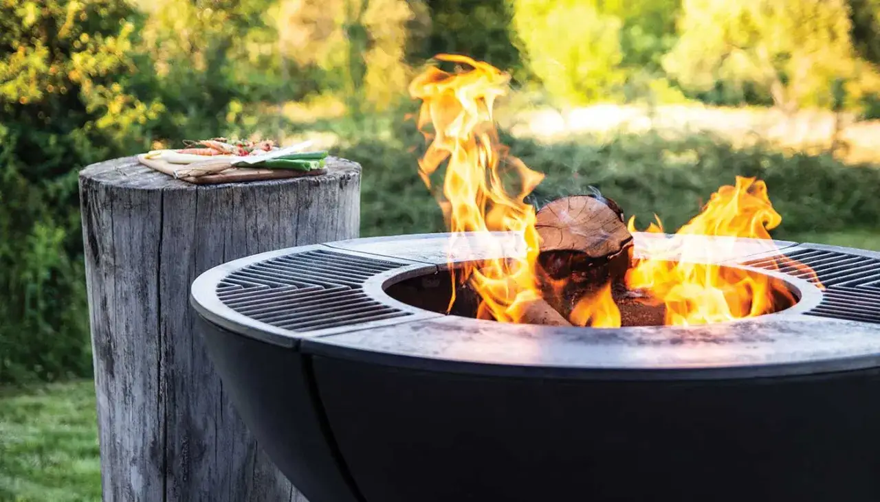 Braciere acceso in giardino, con legna che arde e fiamme vivaci. Sul ceppo di legno, verdure e gamberi pronti per la griglia. Idee giardino con braciere per serate conviviali.