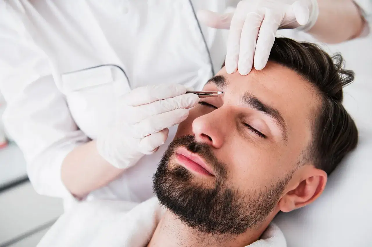 Un homme se fait &eacute;piler le sourcil qui rebique par une esth&eacute;ticienne en blouse blanche et gants.