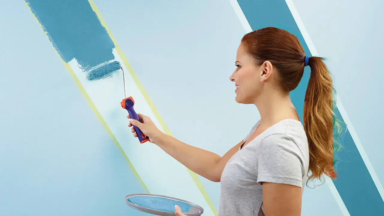 Frau streicht mit einer Farbrolle eine Wand blau. Die Frage, wie oft kann man eine Wand streichen, ist hier nicht relevant, da sie gerade erst beginnt.