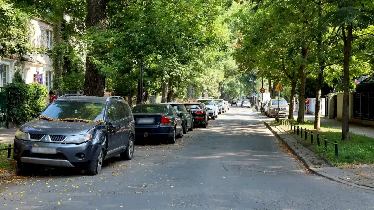 Ulica z zaparkowanymi samochodami, jeden z nich, ciemnoszary SUV, blokuje drogę dojazdową.