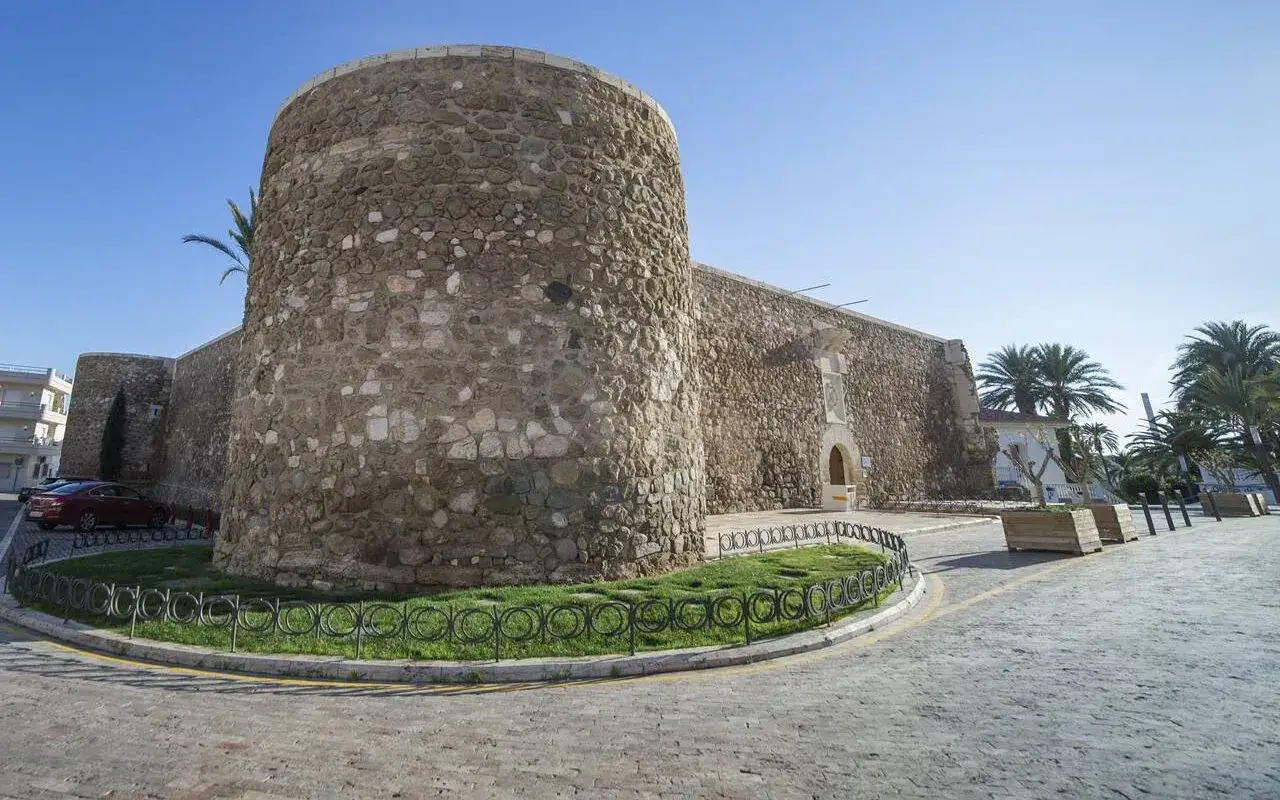 Antigua fortaleza de piedra con torreón circular, situada en el **carboneras paseo marítimo**. Palmeras y cielo azul.