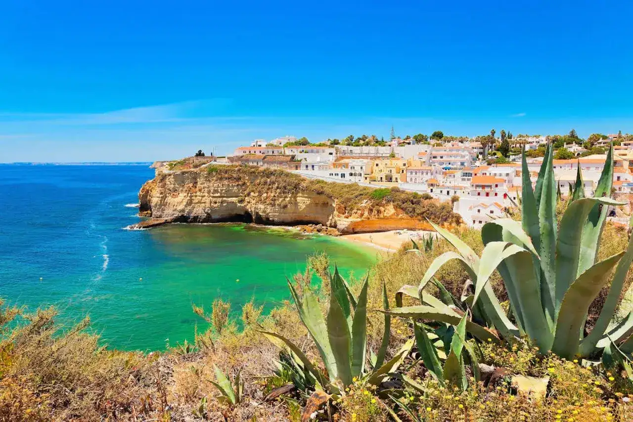 Vue d'une crique aux eaux turquoise, bord&eacute;e de falaises et d'un village blanc, id&eacute;ale pour un camping naturiste Portugal.