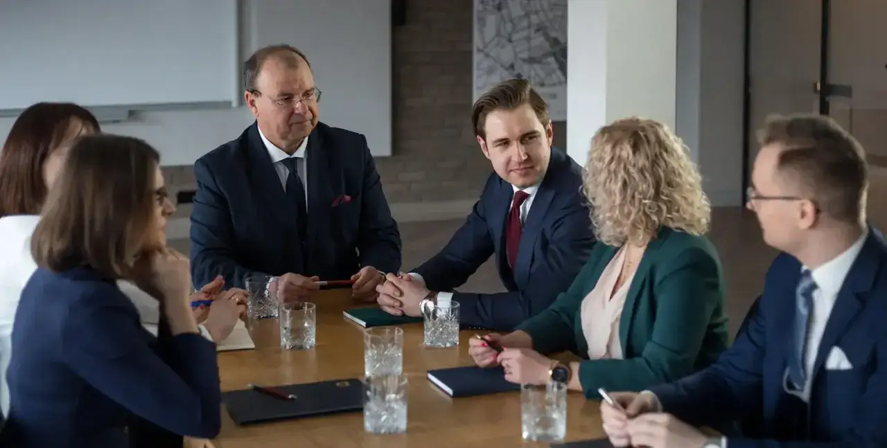 Grupa prawników w garniturach siedzi przy drewnianym stole podczas spotkania.
