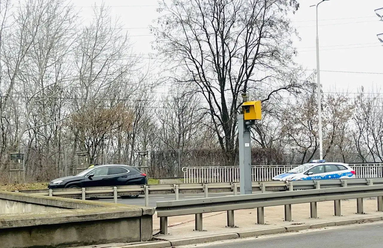 Żółty fotoradar na słupie monitoruje ruch. Obok przejeżdża czarny samochód i radiowóz policyjny. Tak działa fotoradar.