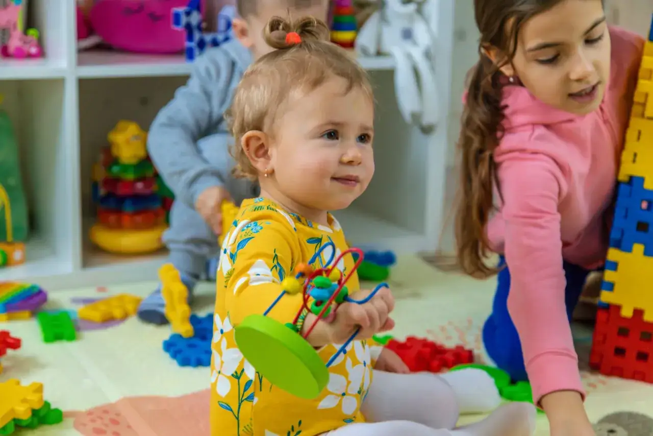 Un bebé de 3 meses con un juguete de cuentas de colores. A su lado, una niña mayor juega con bloques de construcción.