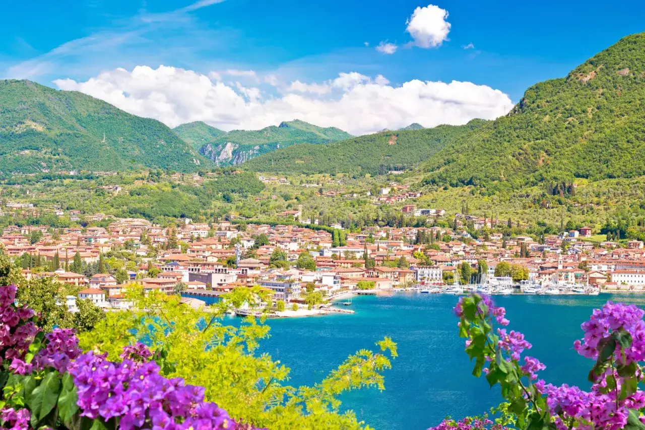 Salò, cosa vedere: un panorama mozzafiato sul lago con montagne verdi, case colorate e fiori viola in primo piano.