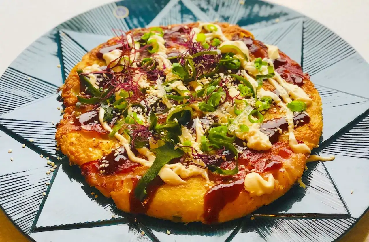 En läcker japansk kålpannkaka toppad med majonnäs, salladslök och sesamfrön på en blåmönstrad tallrik.