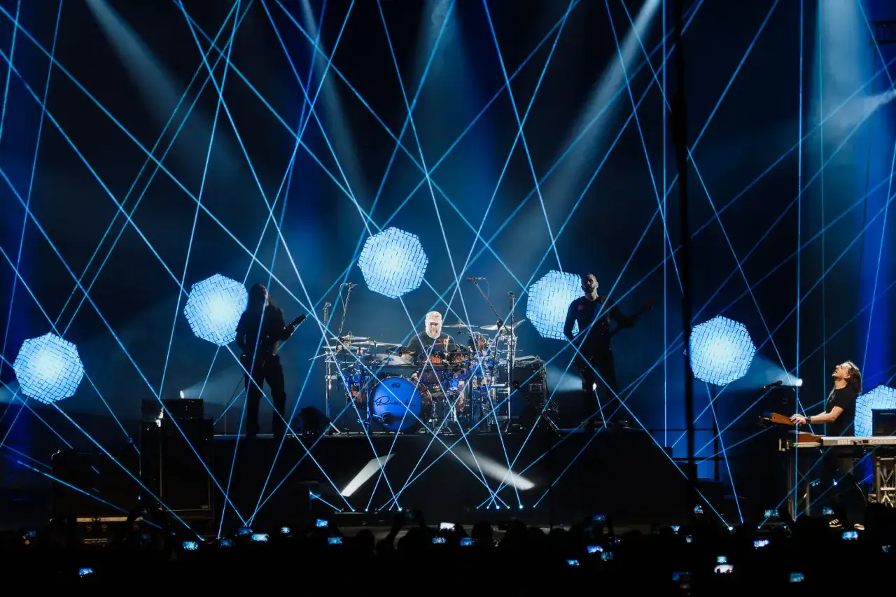 Zespół gra na scenie, otoczony niebieskimi laserami. Perkusista siedzi za zestawem, gitarzysta gra, a klawiszowiec tworzy muzykę. Koncert odbywa się nad rzeką, a publiczność filmuje występy.