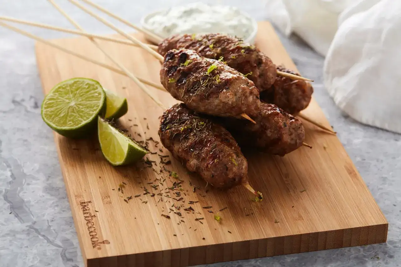 Brochettes d'agneau grillées, garnies d'herbes et de zeste de citron vert, servies avec des quartiers de citron vert sur une planche en bois.