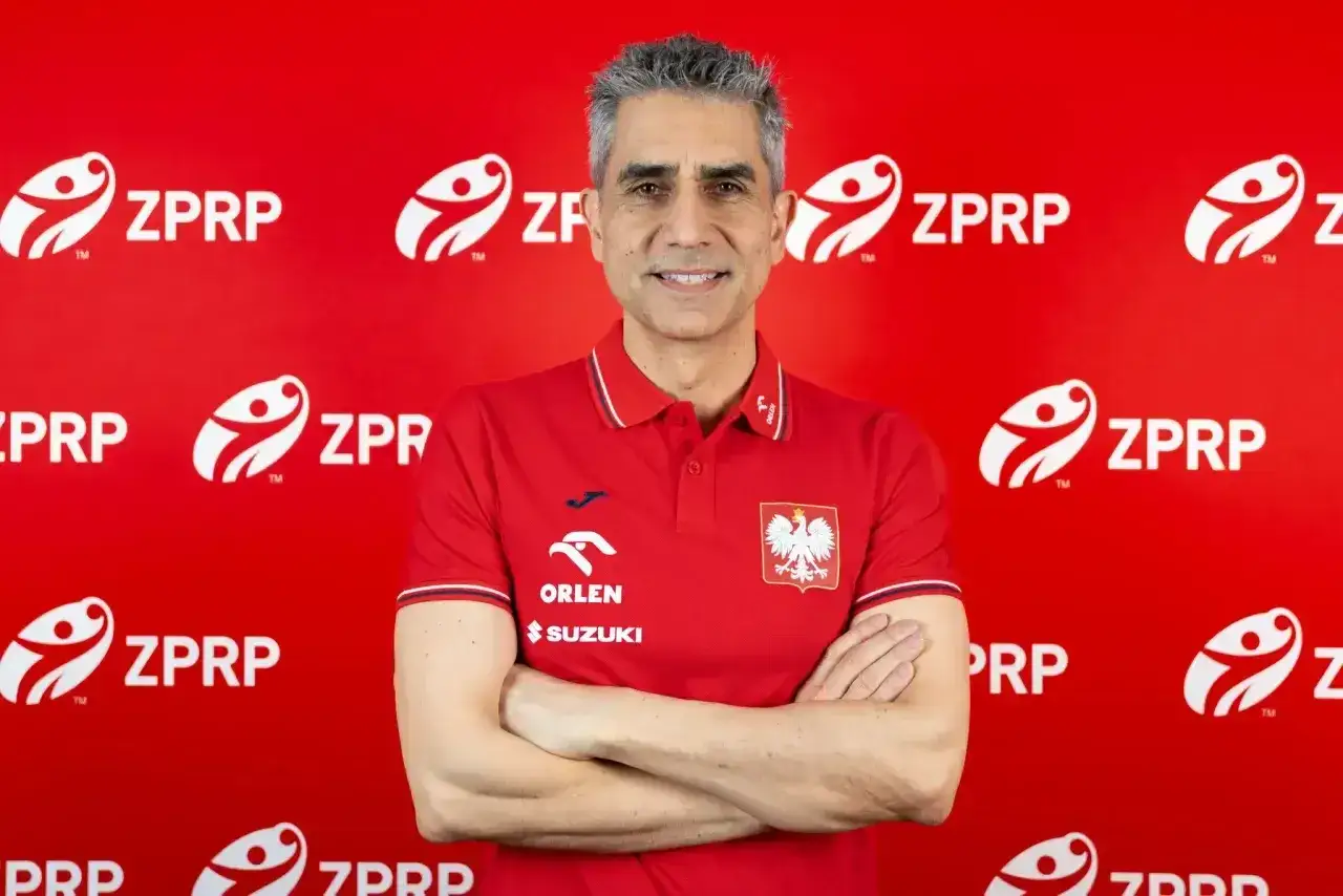 Uśmiechnięty mężczyzna w czerwonej koszulce z godłem Polski, były trener piłkarzy ręcznych, stoi na tle czerwonej ściany z logo ZPRP.