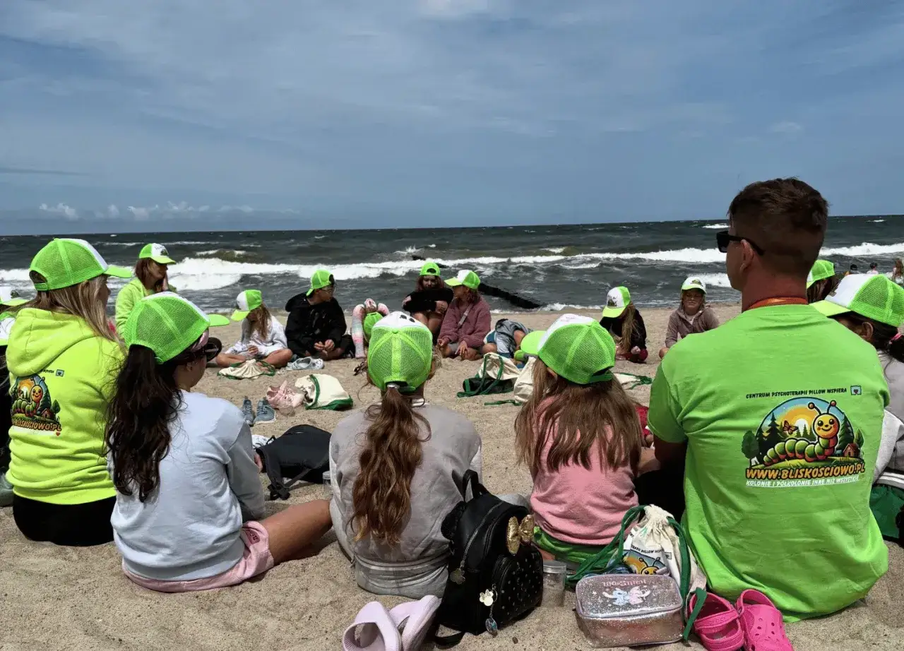 Dzieci w zielonych czapkach siedzą na plaży, słuchając opiekuna. To z pewnością sprawdzone kolonie dla dzieci, gdzie nauka łączy się z zabawą nad morzem.