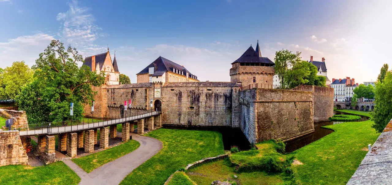 Le **Château de Nantes** se dresse fièrement, entouré de douves verdoyantes. Un pont mène à son entrée fortifiée, sous un ciel bleu clair.