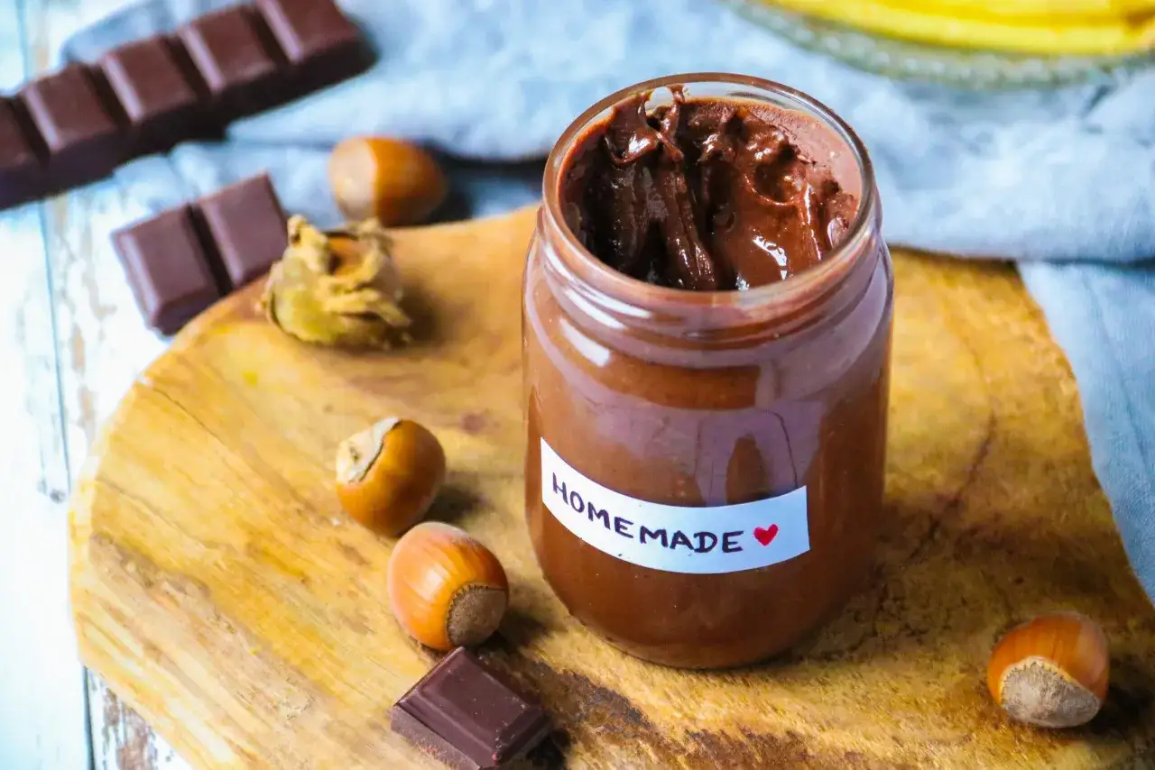 Pâte à tartiner maison, onctueuse et chocolatée, rappelant le goût du Kinder Bueno. Des noisettes et des carrés de chocolat complètent la scène gourmande.