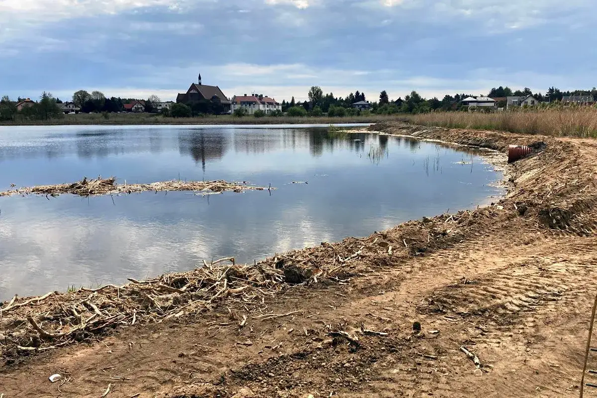 Zalew Zielona Kaleta, spokojna tafla wody odbija chmury. Na brzegu widać ślady prac ziemnych i pozostałości roślinności. W oddali kości&oacute;ł i zabudowania.
