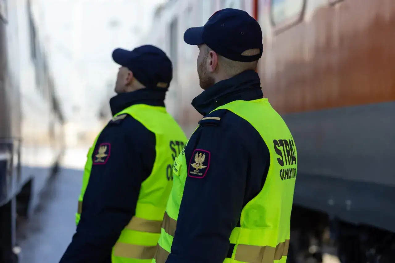 Dwóch funkcjonariuszy Służby Ochrony Kolei w kamizelkach odblaskowych stoi przy pociągu. Praca w SOK to odpowiedzialność.