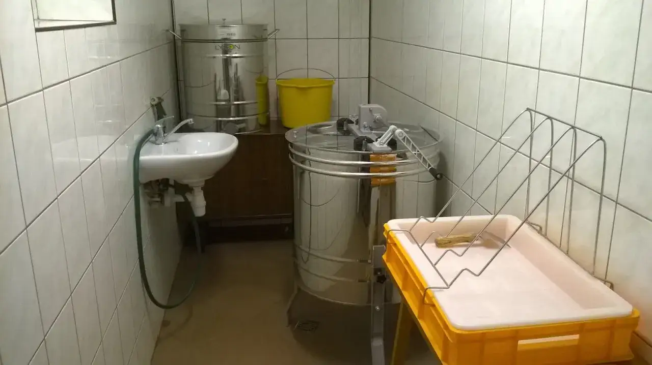W pomieszczeniu pszczelarza stoi miodarka, beczki na miód, żółte wiadro i zlew.