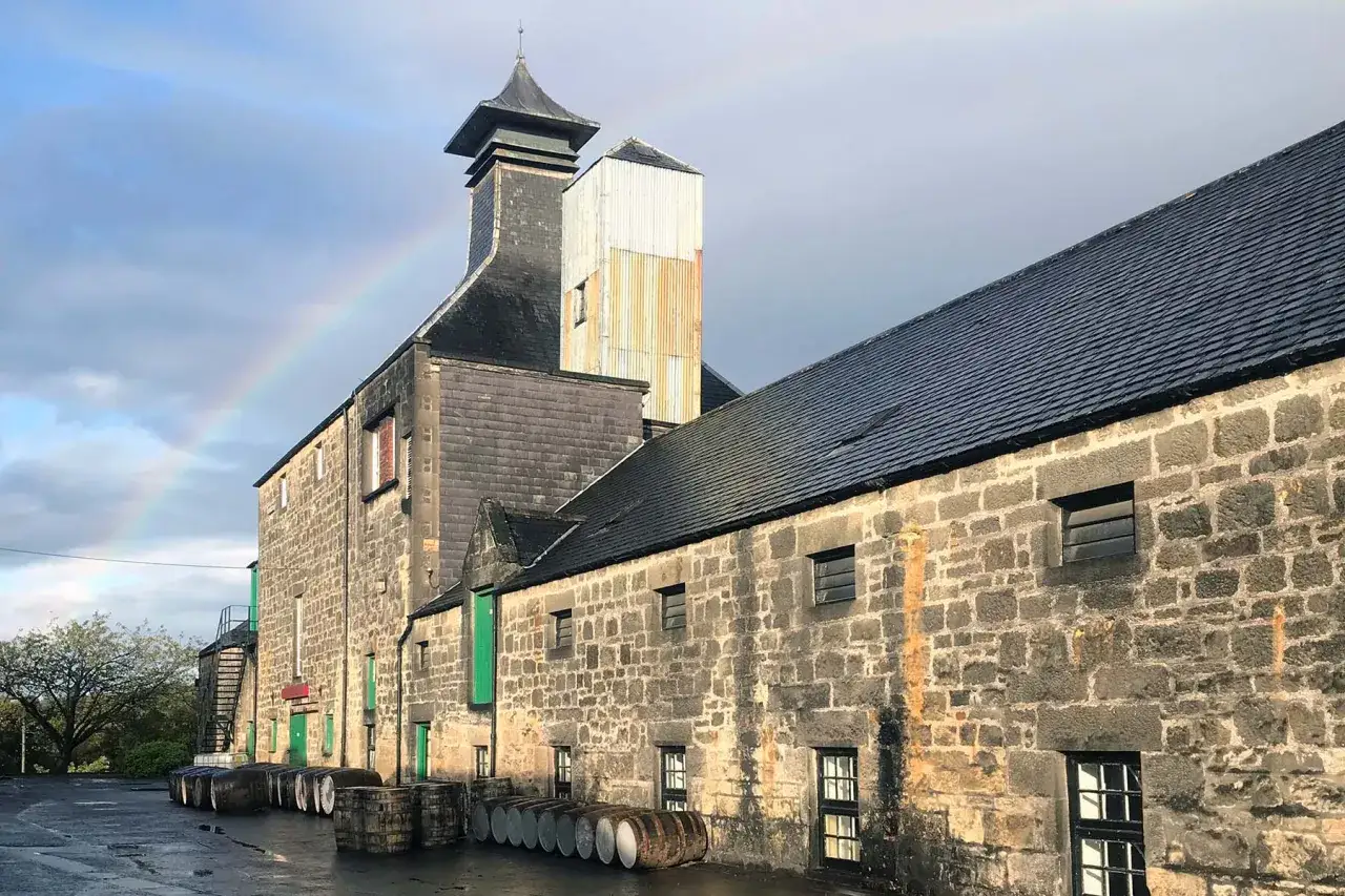 Un arc-en-ciel apparaît au-dessus de la distillerie Benriach, avec des tonneaux de whisky alignés devant les bâtiments en pierre.