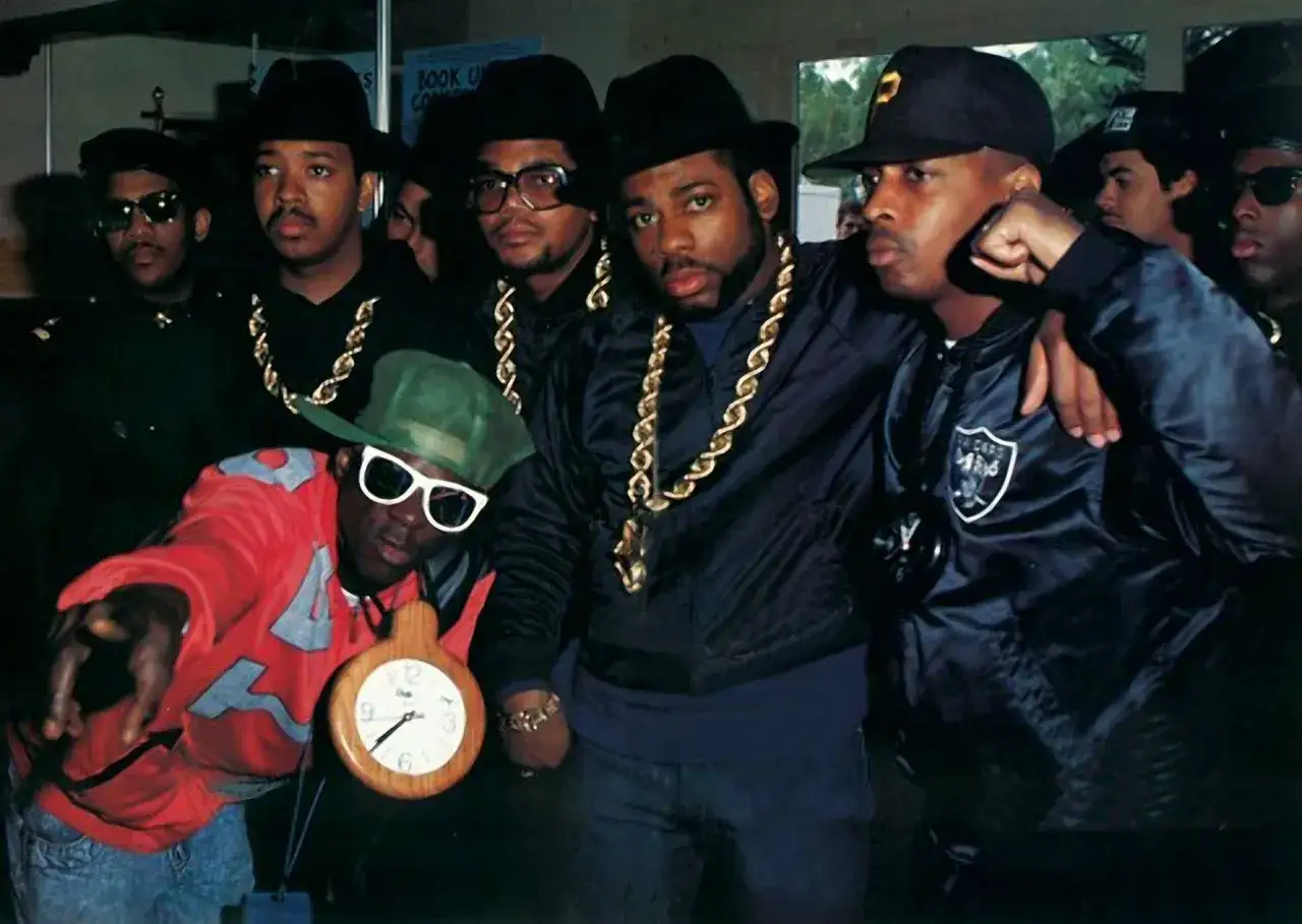 Un groupe de rappeurs années 80, arborant chaînes en or, chapeaux et vestes stylées, pose pour la caméra.