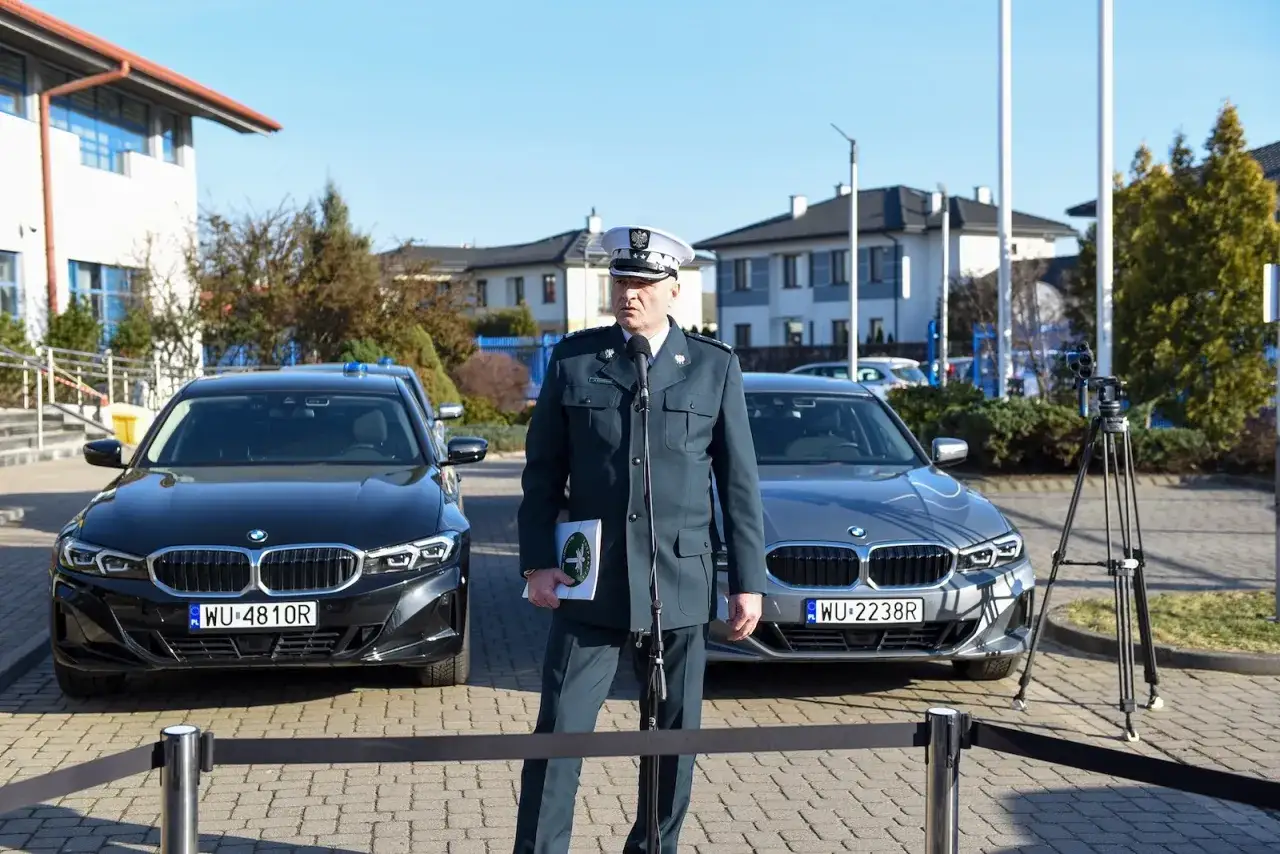 Funkcjonariusz w mundurze stoi przed dwoma BMW, jednym czarnym, drugim szarym. To nieoznakowany radiowóz, gotowy do akcji.