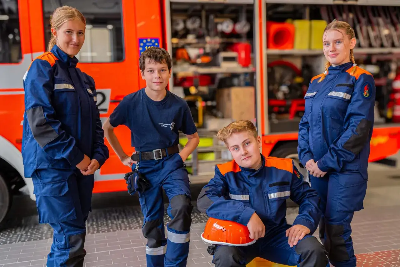 Vier junge Leute in blauer Einsatzkleidung stehen vor einem Feuerwehrauto. Die Jugendfeuerwehr ist bereit f&uuml;r ihren Einsatz.